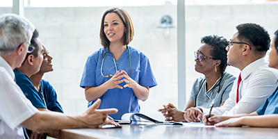 a woman wearing a stethoscope speaks people sitting around a table